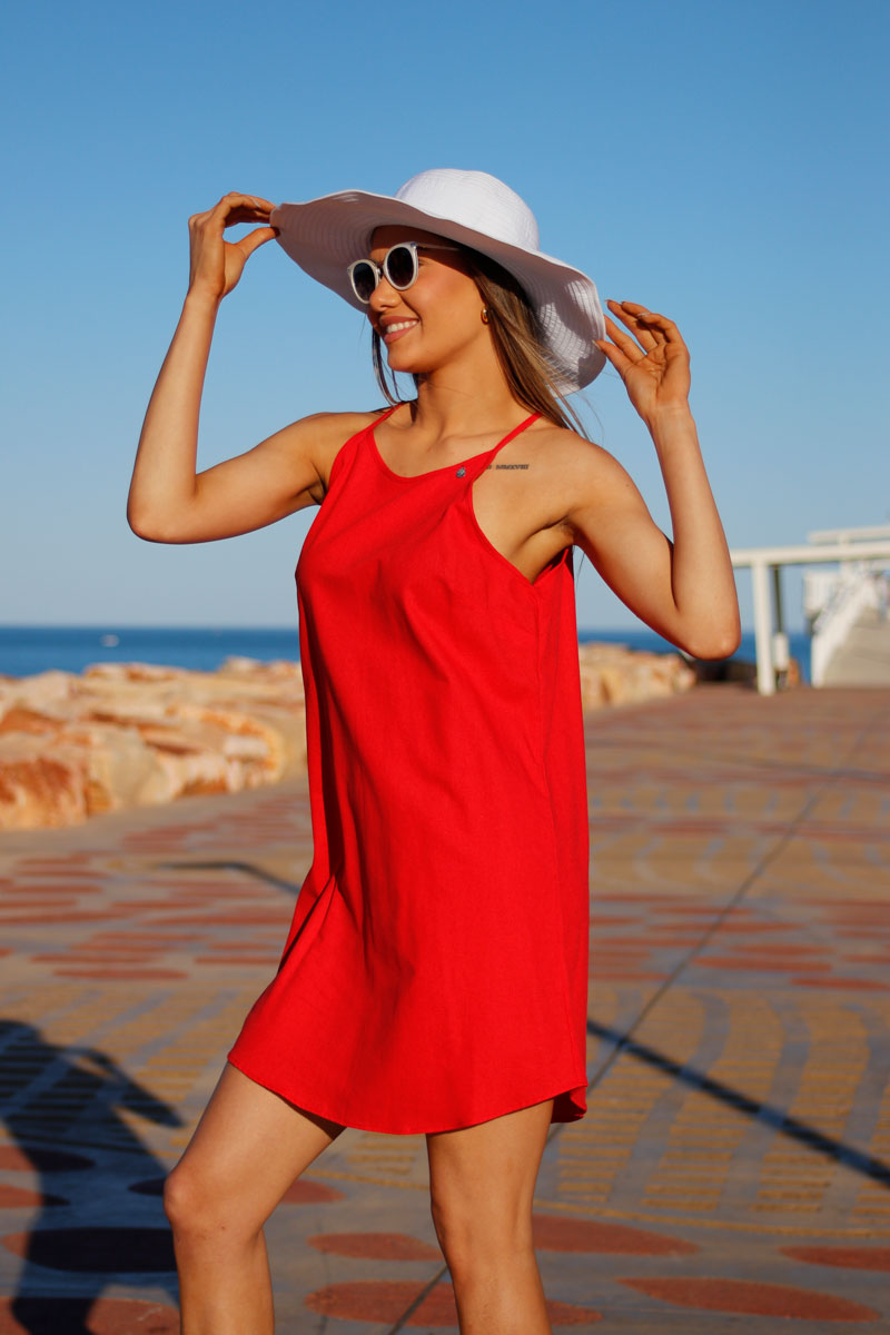 A woman in a red sleeveless knee-length linen dress, wearing sunglasses and a wide-brimmed hat, standing by the sea.