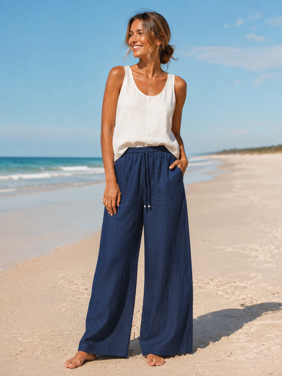 A woman stands on a sandy beach wearing loose navy wide-leg pants and a white sleeveless top, with the ocean and blue sky in the background.